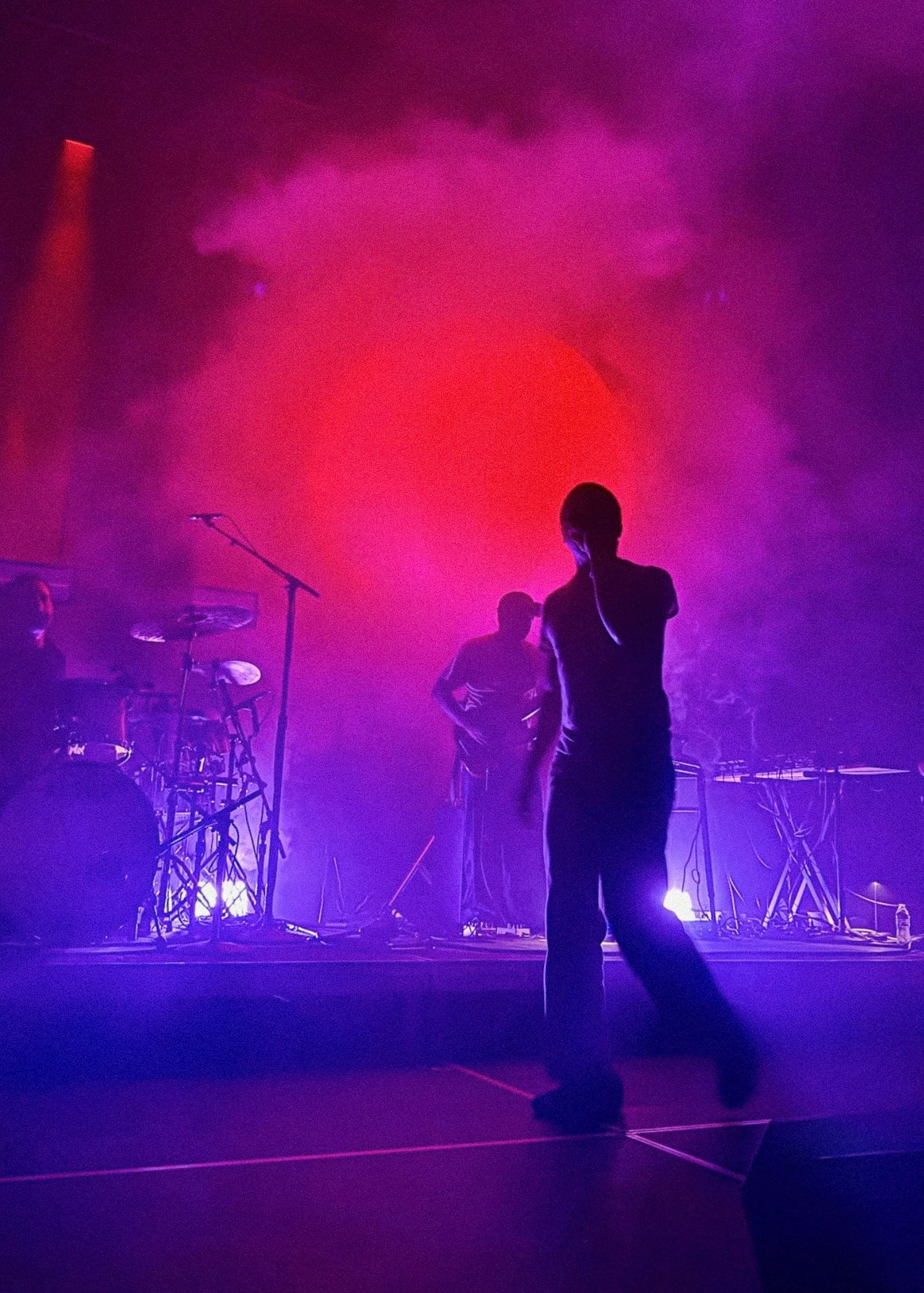 A figure silhouetted in magenta haze in front of the band, the stage washed pink and purple