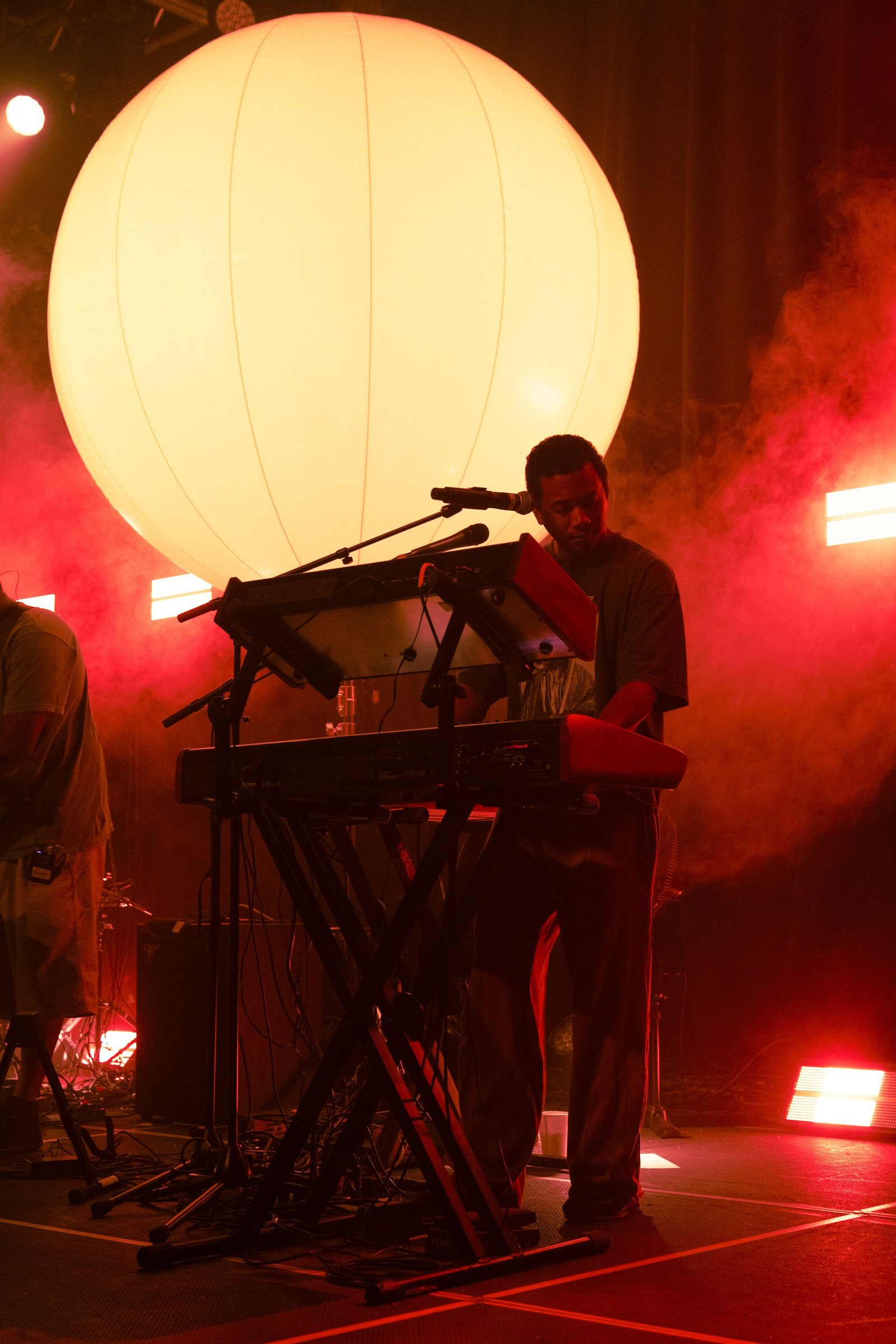 Ball on Toro y Moi's 2026 tour — Chaz Bear at the keys with the six-foot LED sphere glowing behind him through red haze