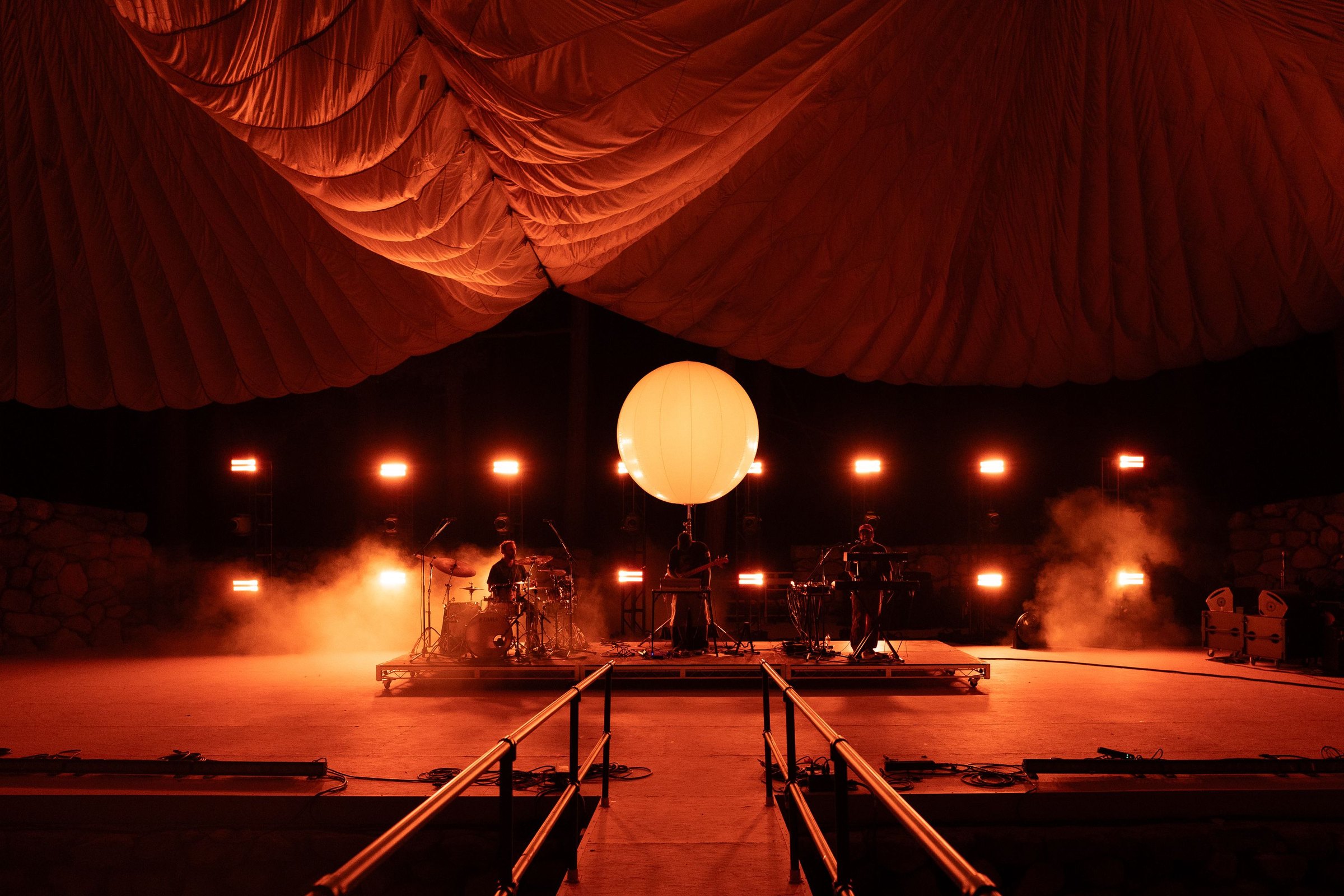 The full stage seen head-on under the tent roof, the sphere centred between banks of strobes with fog across the empty floor