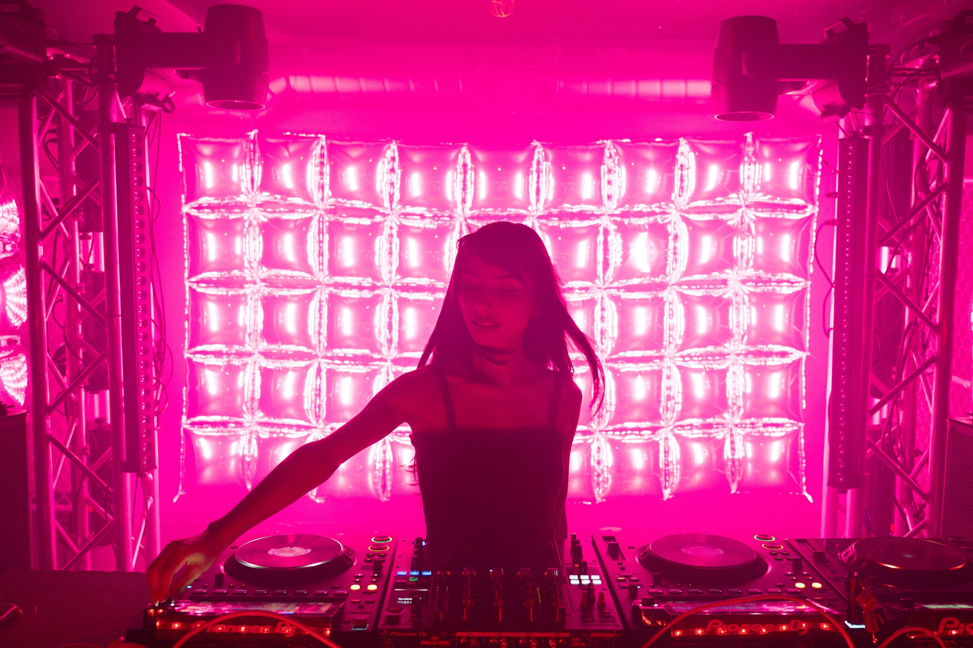 Erika silhouetted at the decks against the balloon wall glowing hot pink, truss towers standing on either side