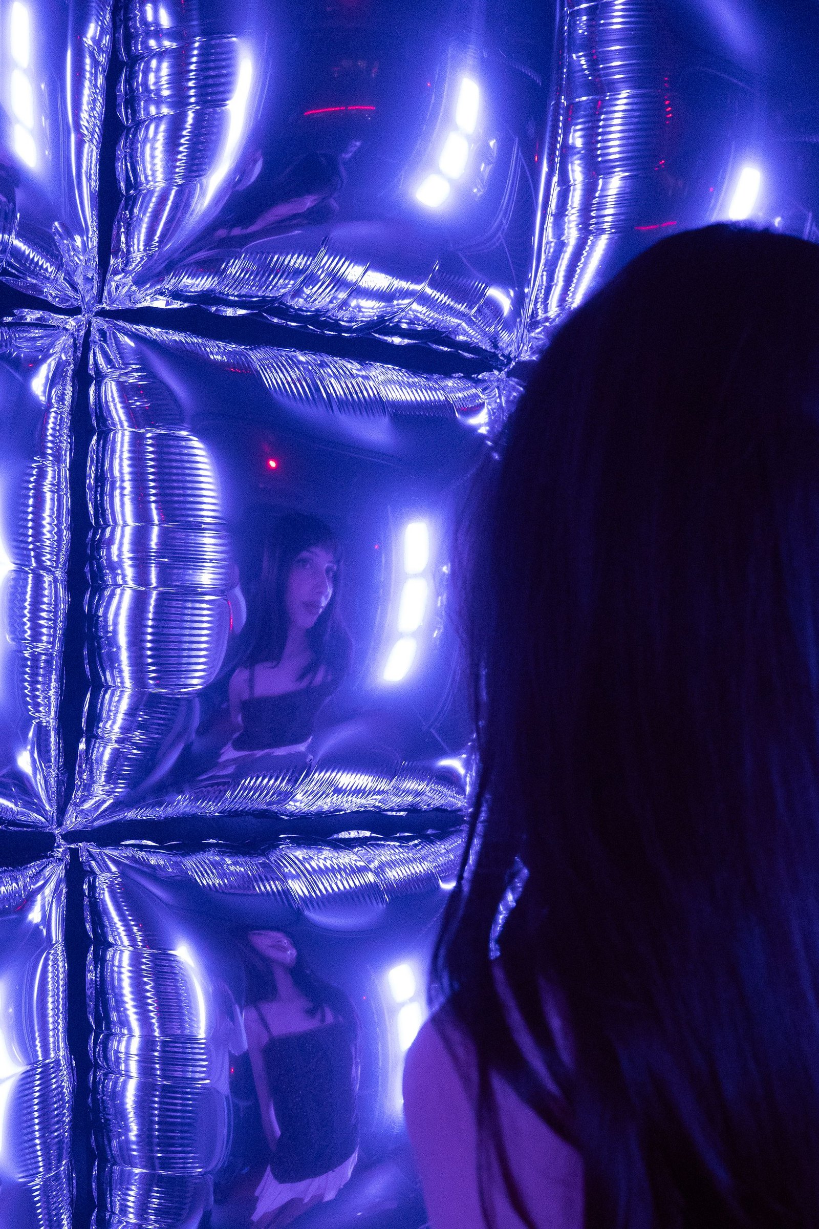 Erika looking into the chrome balloon wall lit blue, her reflection repeated across the curved mylar surfaces