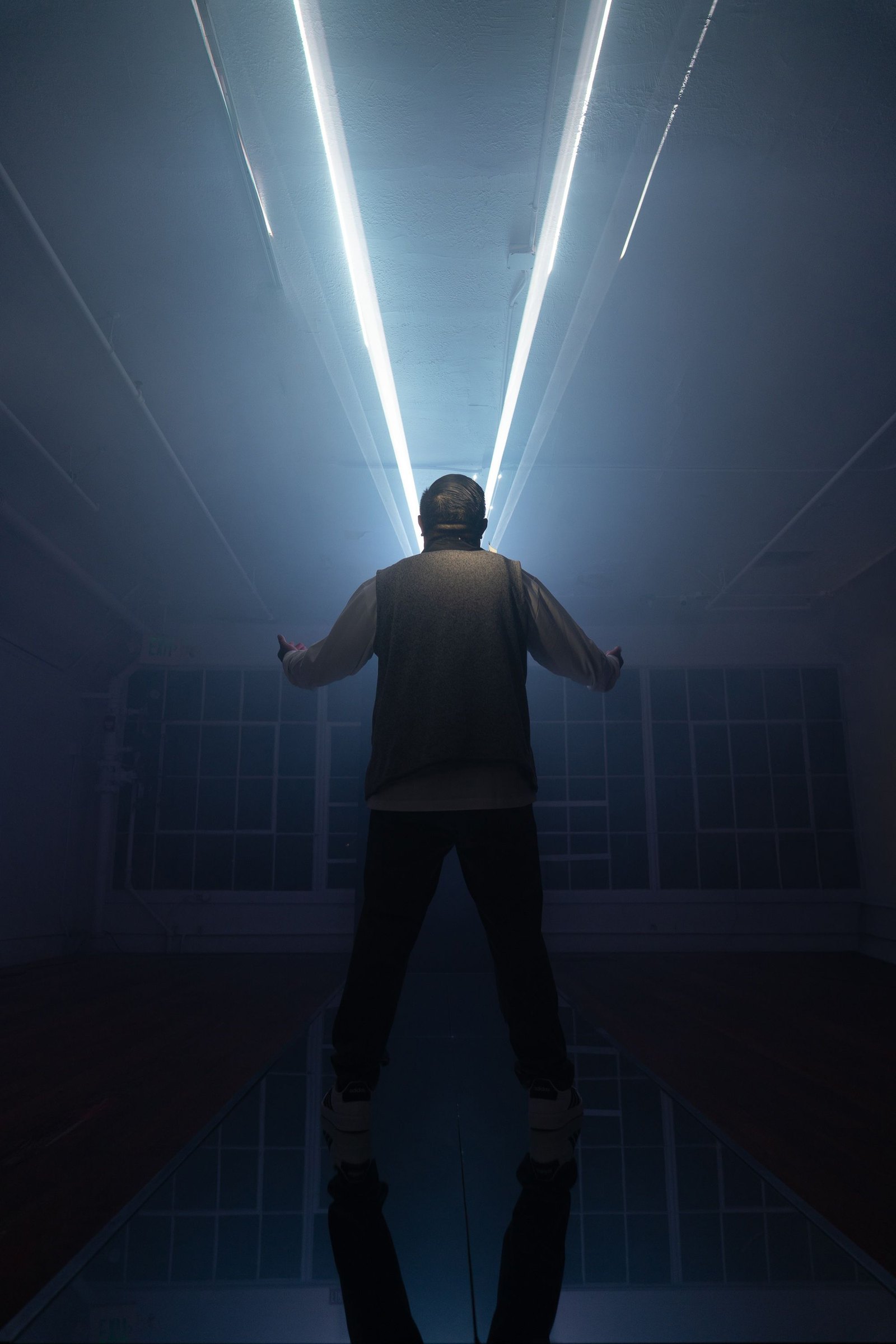 A man seen from behind with his arms held out, standing on the mirrored catwalk under two white laser beams that run the length of the ceiling