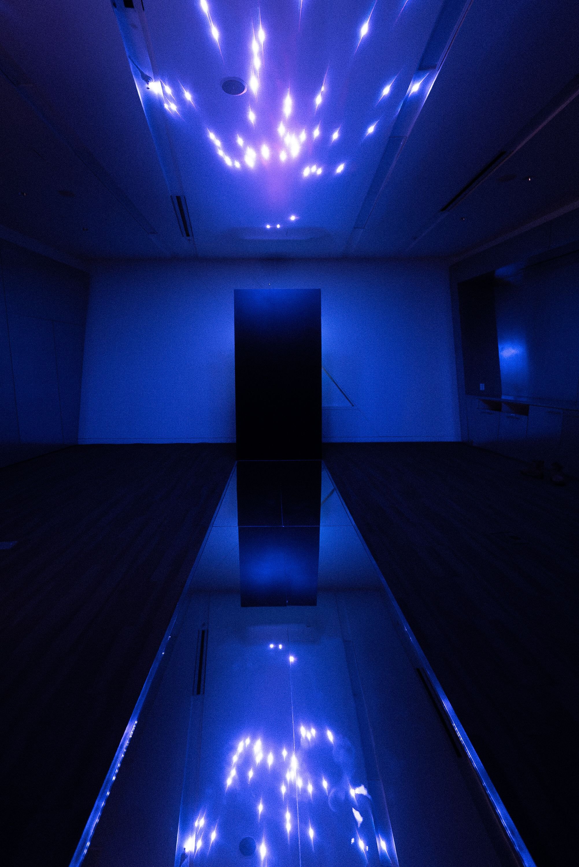 Looking down the mirrored catwalk to the black acrylic column, blue-violet laser points scattered across the ceiling and doubled in the mirror, at the Contemporary Jewish Museum