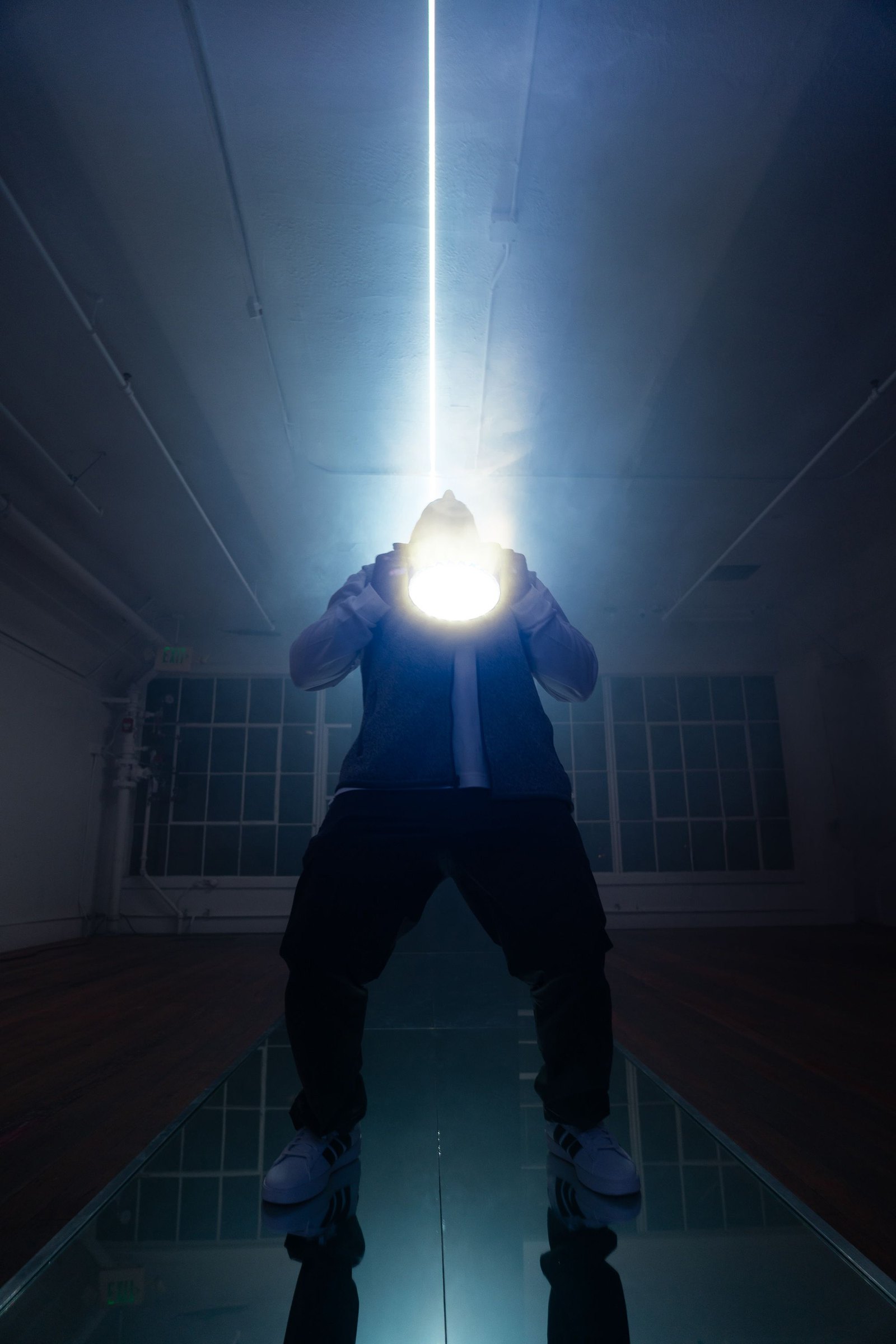 A figure on the mirrored catwalk holding a blinding white light at their face, a single laser beam rising from it to the ceiling