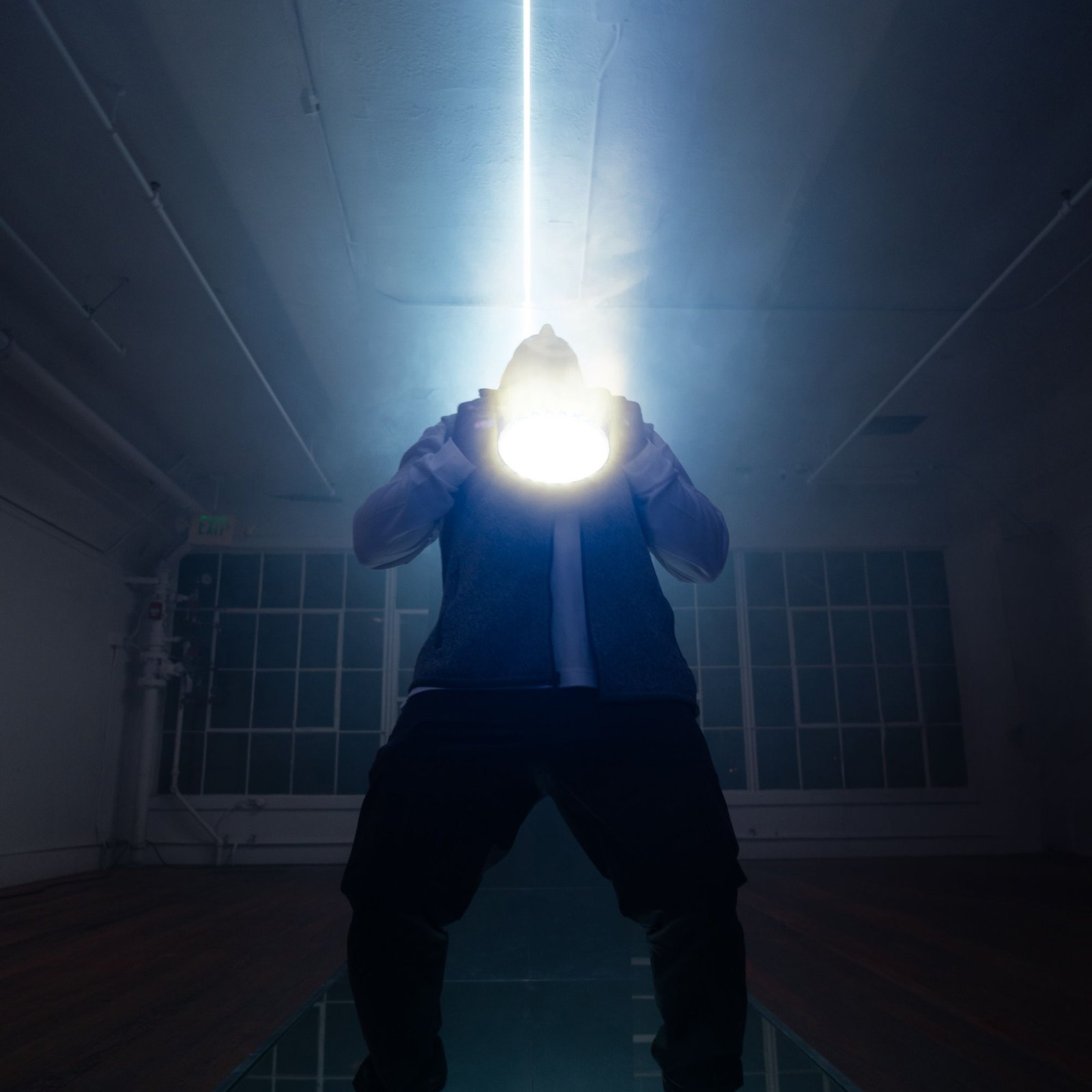 Creative Pulse — a figure on the mirrored catwalk holding a blinding white light at their face, a single laser beam rising from it to the ceiling