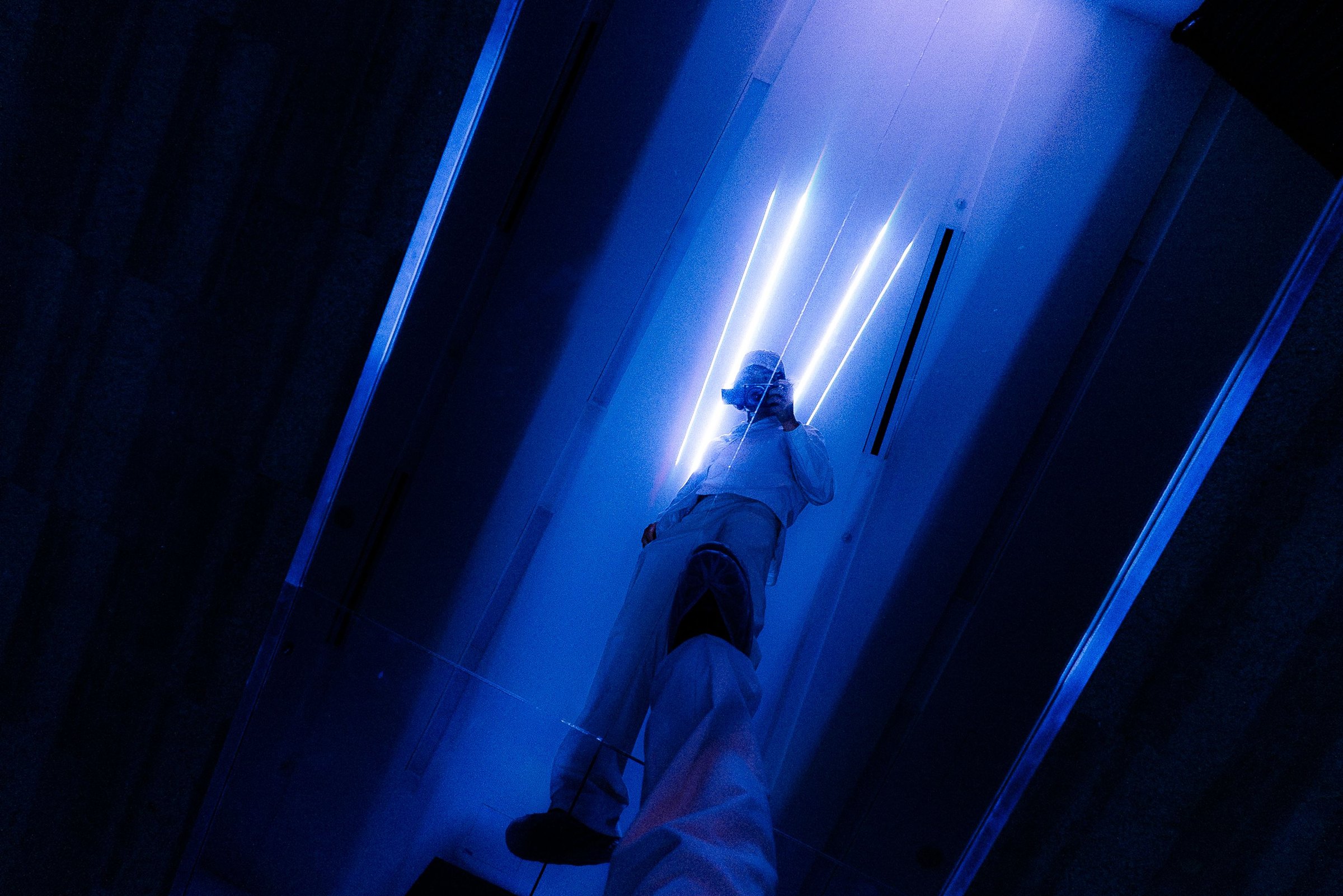 A visitor in white on the mirrored catwalk, seen in its reflection from below, with white laser lines on the ceiling and the aluminum edges glowing blue