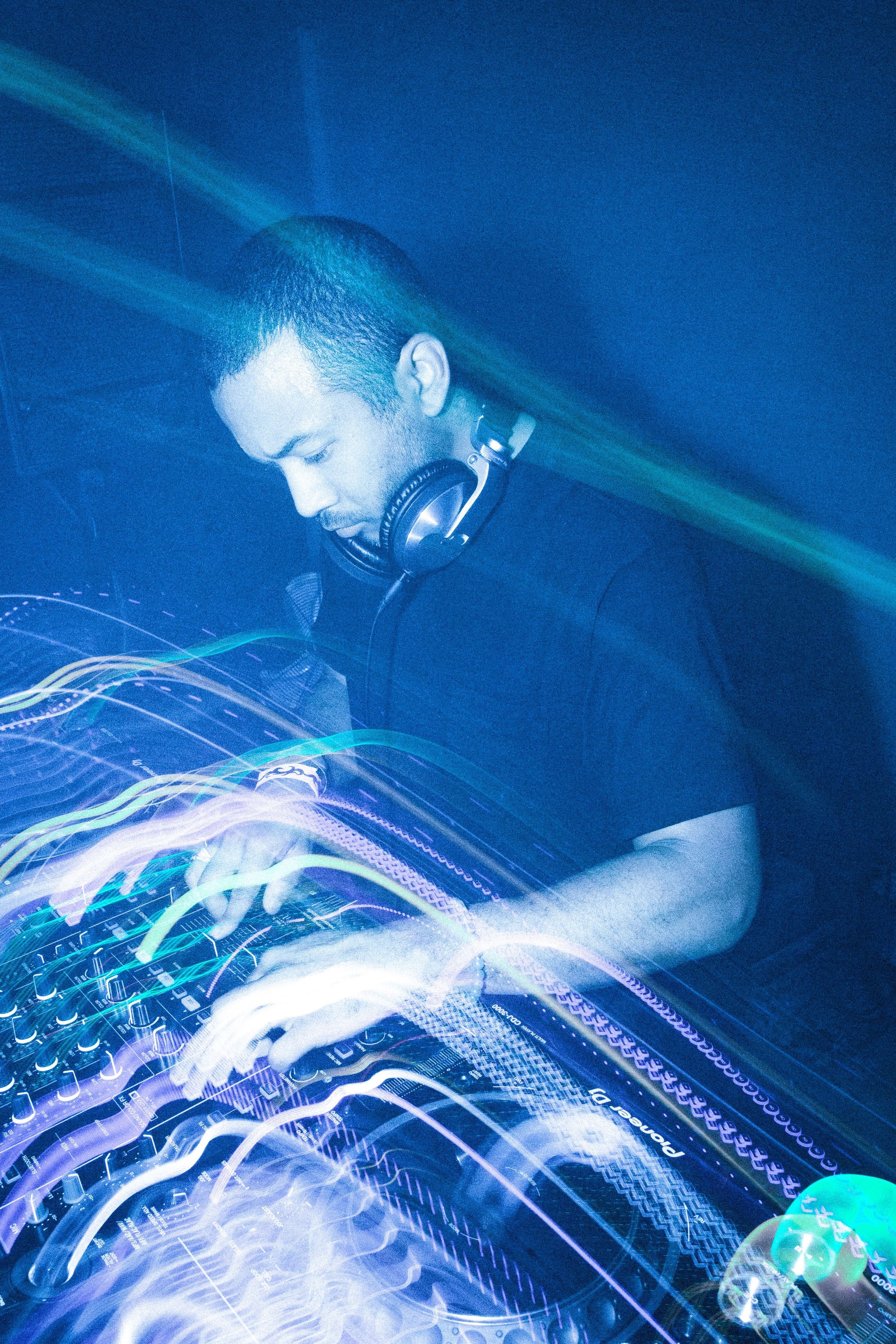 Chazwick Bear at the decks in blue light, long-exposure streaks trailing across the mixer
