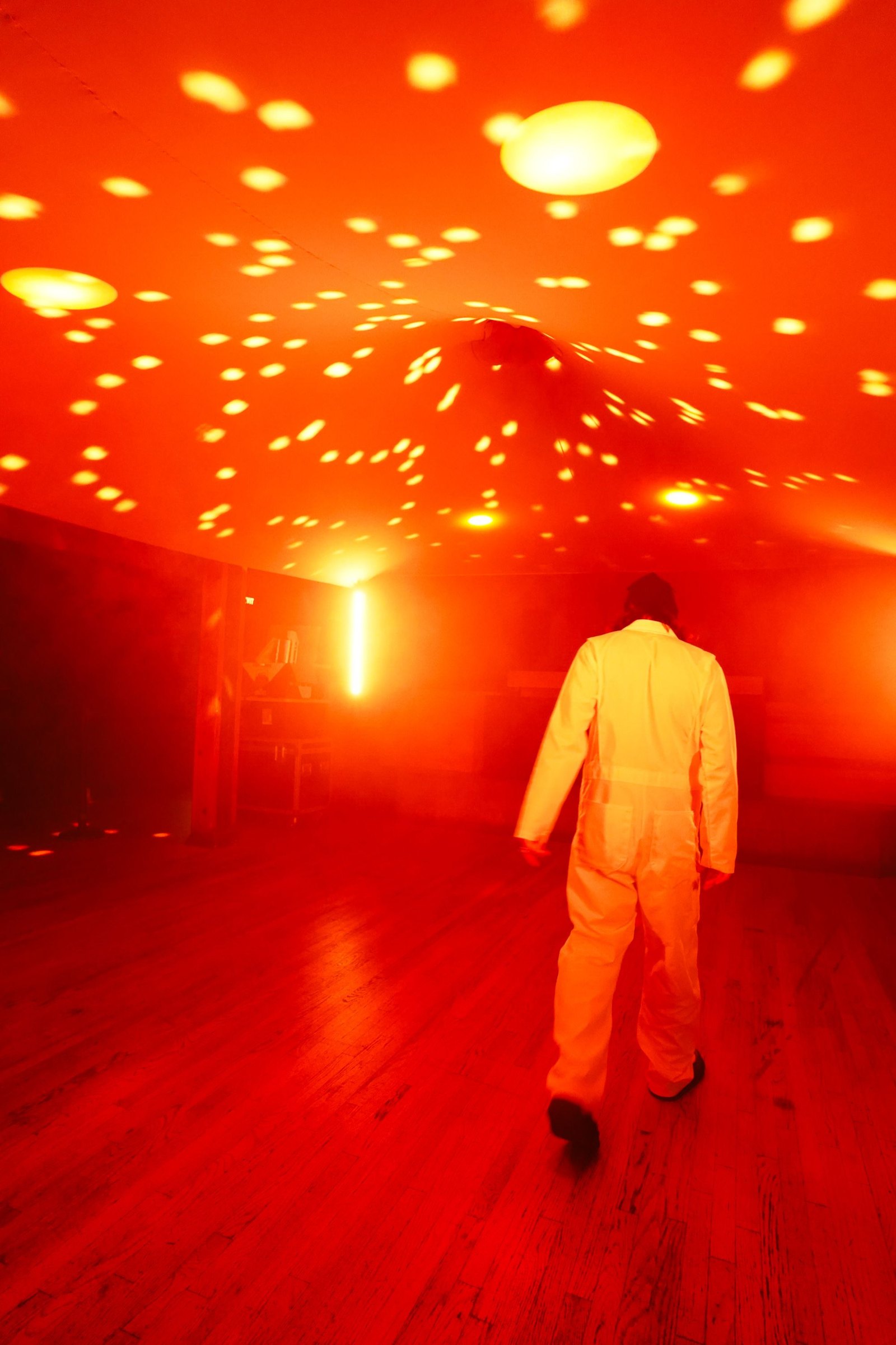 A figure in a white suit walking away beneath the canopy, its fabric speckled with orange light from the disco ball
