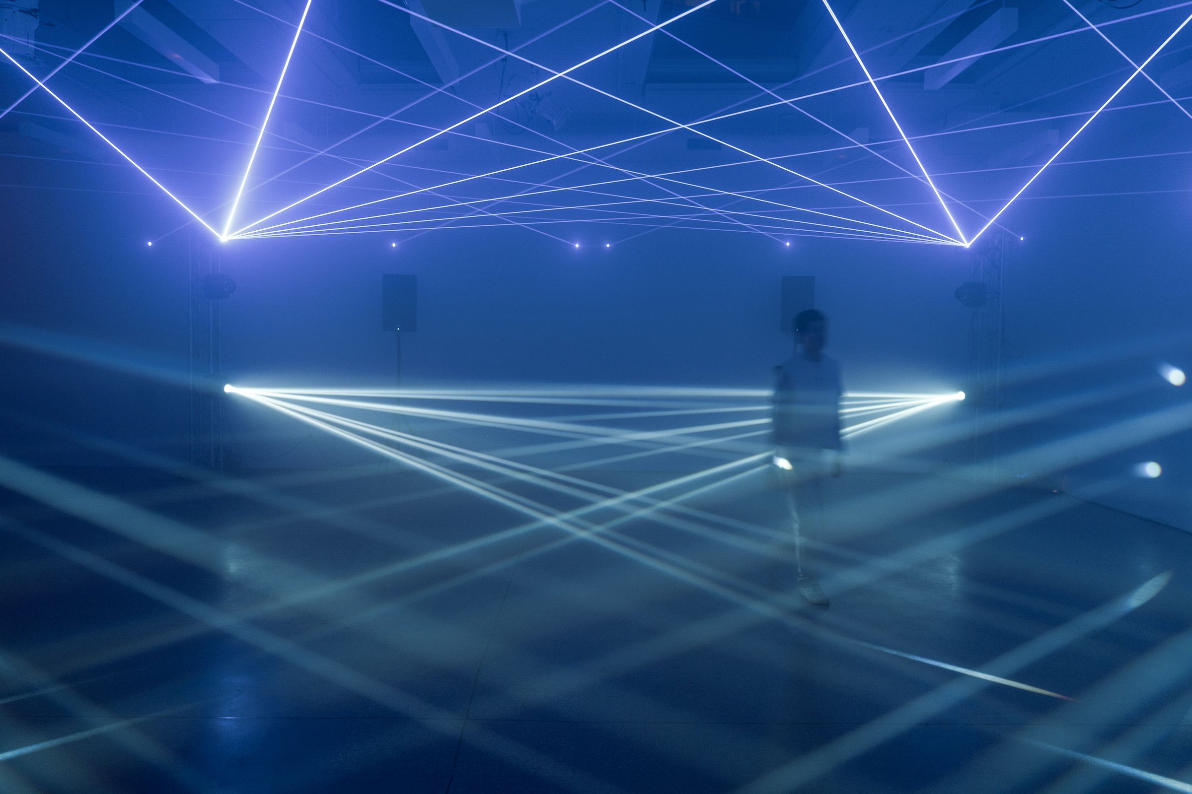 Lattice at Four One Nine — a lone figure in the fog beneath a grid of laser lines crossing overhead and fanning low across the floor