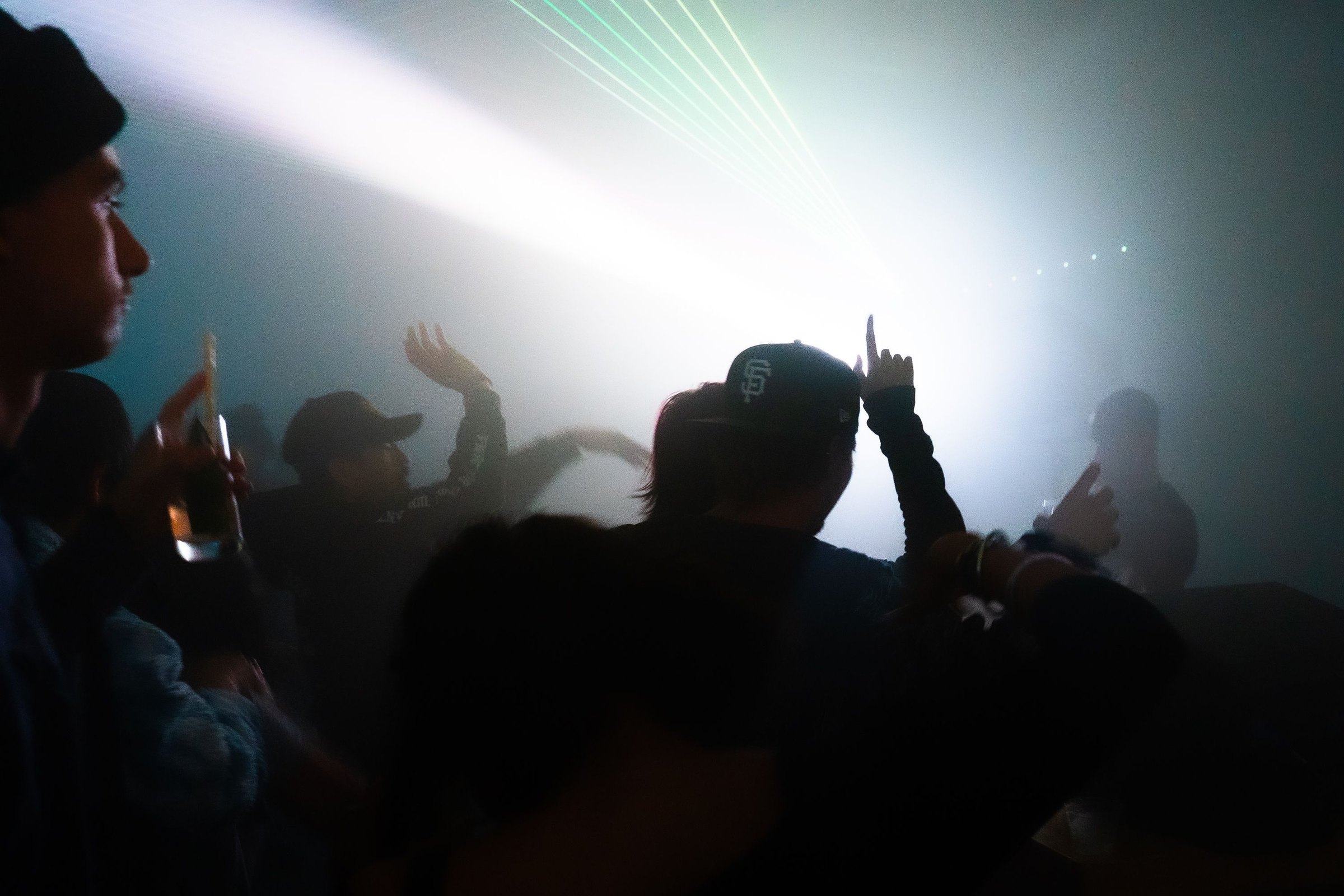 Hands raised in the crowd as a green laser fan cuts through white haze over the dancefloor