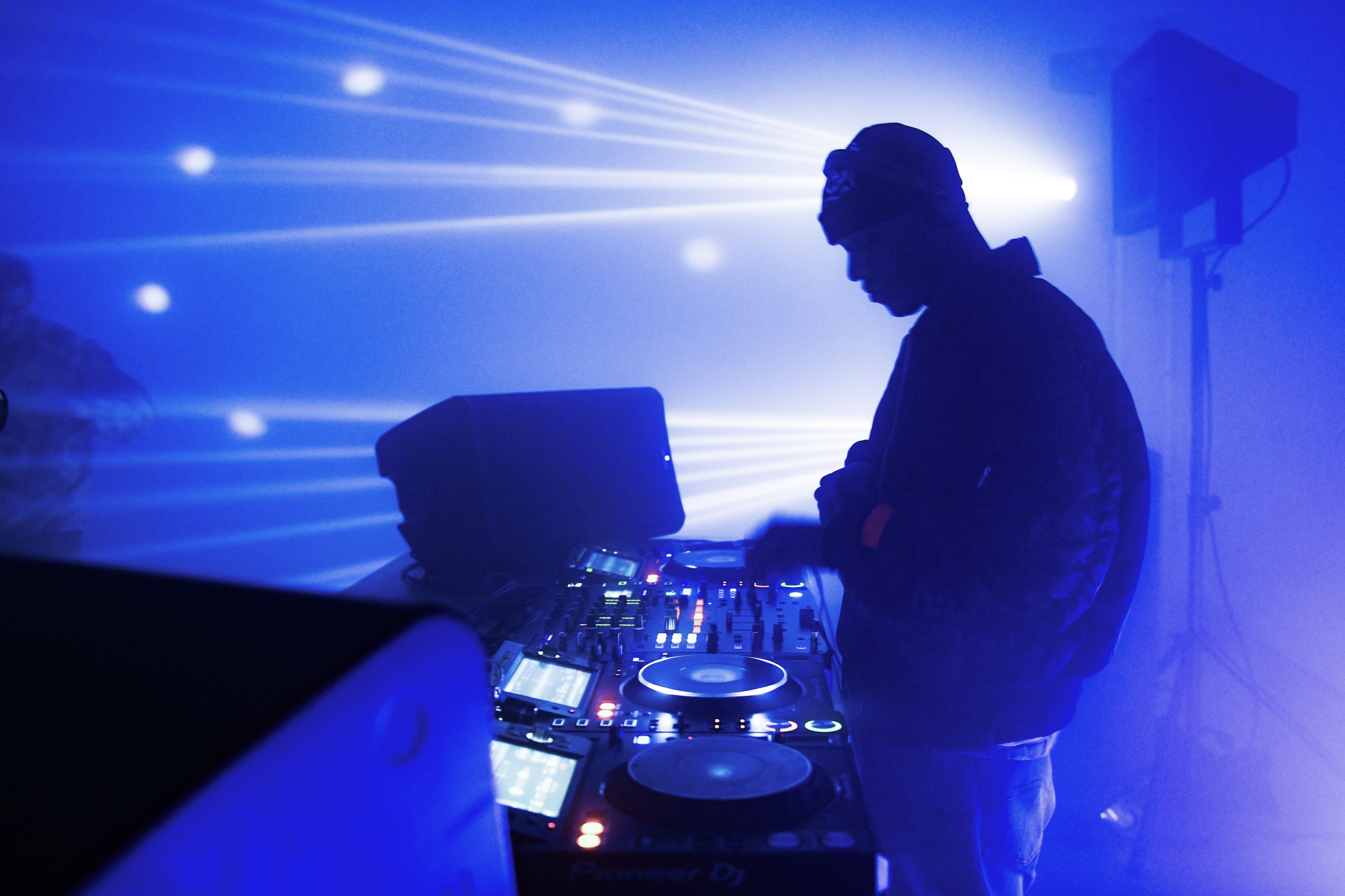 Heron Preston silhouetted at the CDJs, blue laser lines fanning across the wall behind the booth