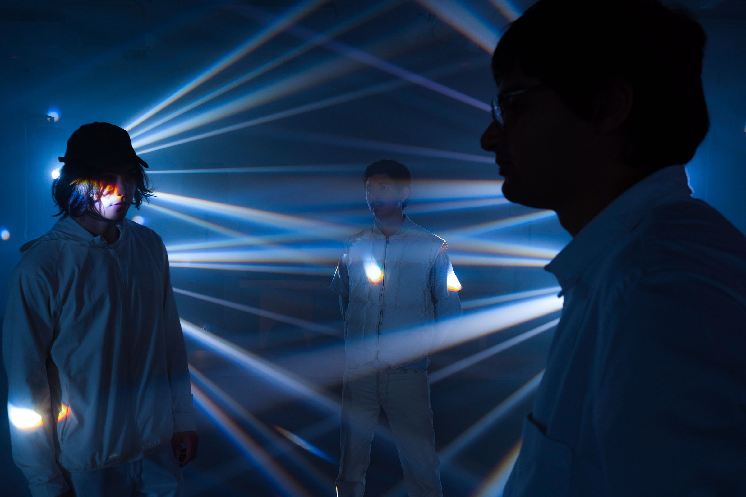 Three New Nostalgia members in white among the laser beams, two in profile and one lit from below at the center of the room