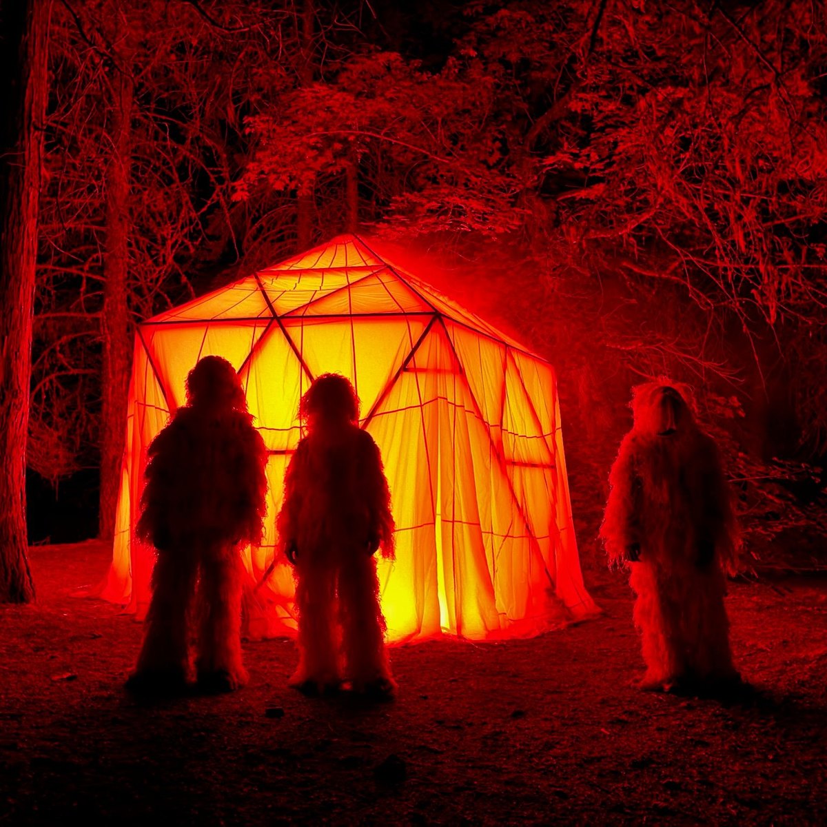 Three New Nostalgia members in white ghillie suits standing before HoloDome3, a parachute-covered dome glowing orange in the forest at night