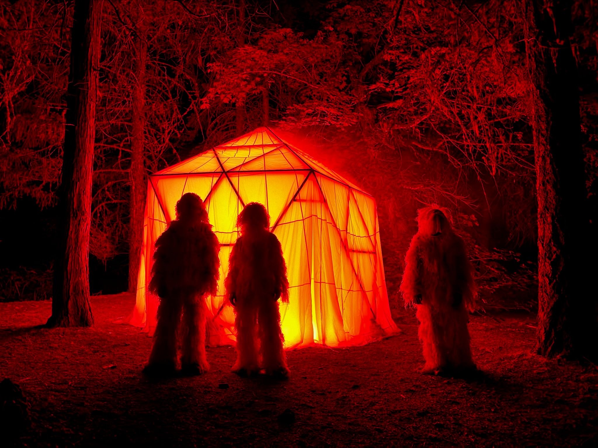 Three New Nostalgia members in white ghillie suits standing before HoloDome3, a parachute-covered dome glowing orange in the forest at night