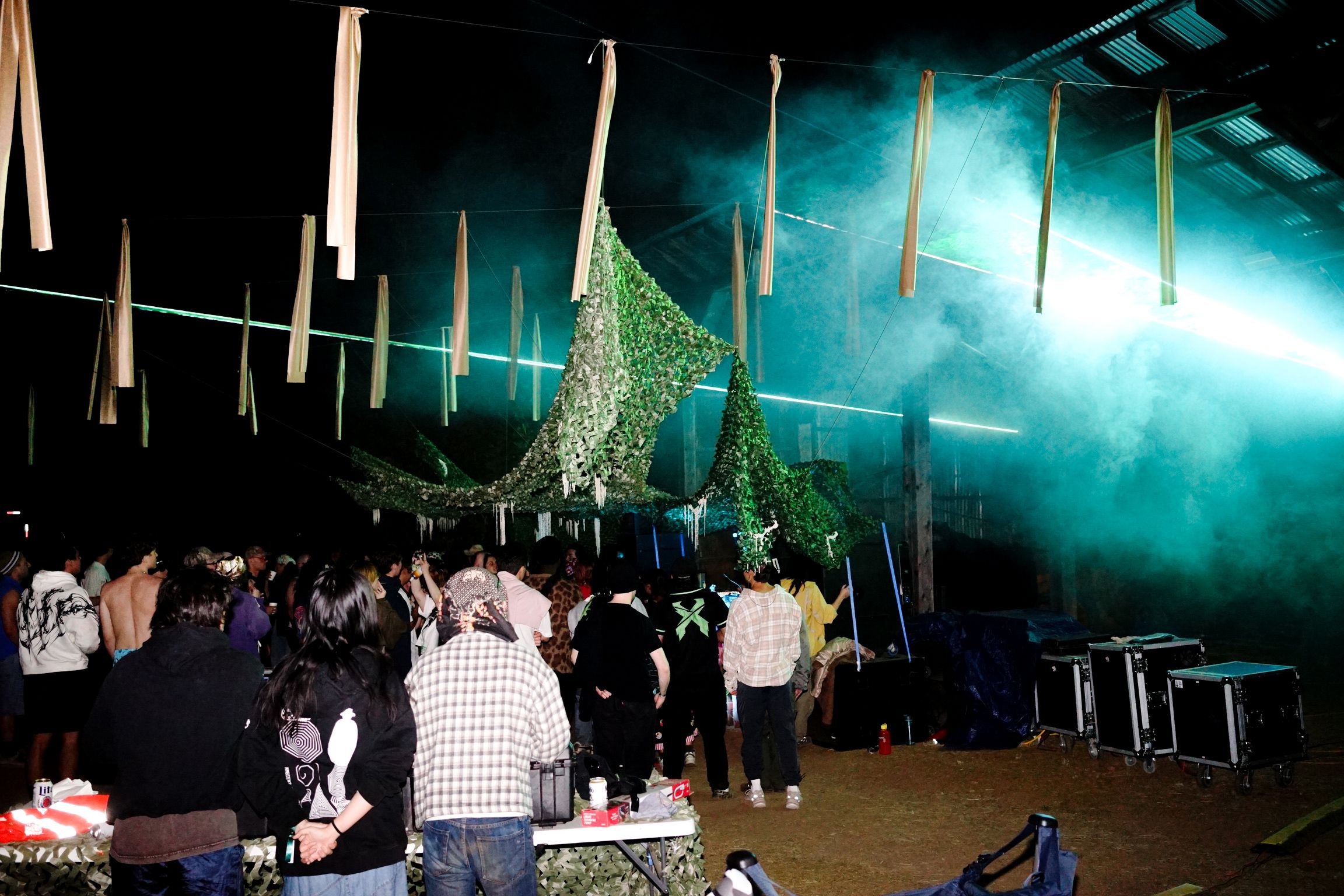 The crowd under camouflage netting at The Club, lasers cutting through fog above the corrugated shed